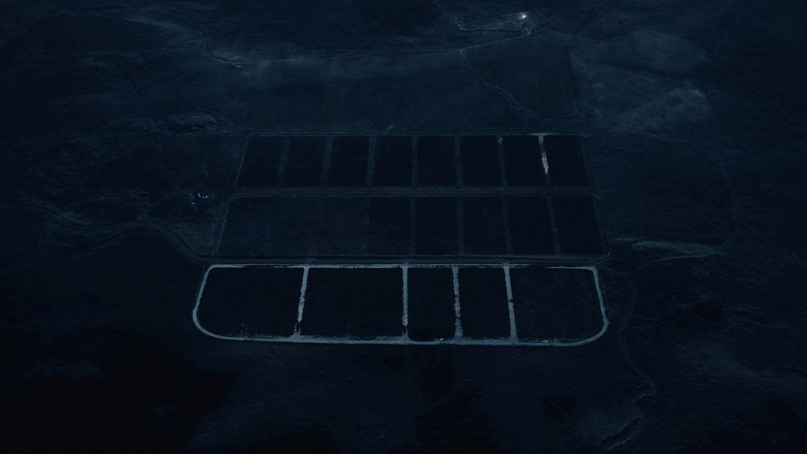 Night aerial looking straight down on an exurban land grid: rows of dark rectangular undeveloped parcels separated by pale gravel roads, with one distant farmyard light.