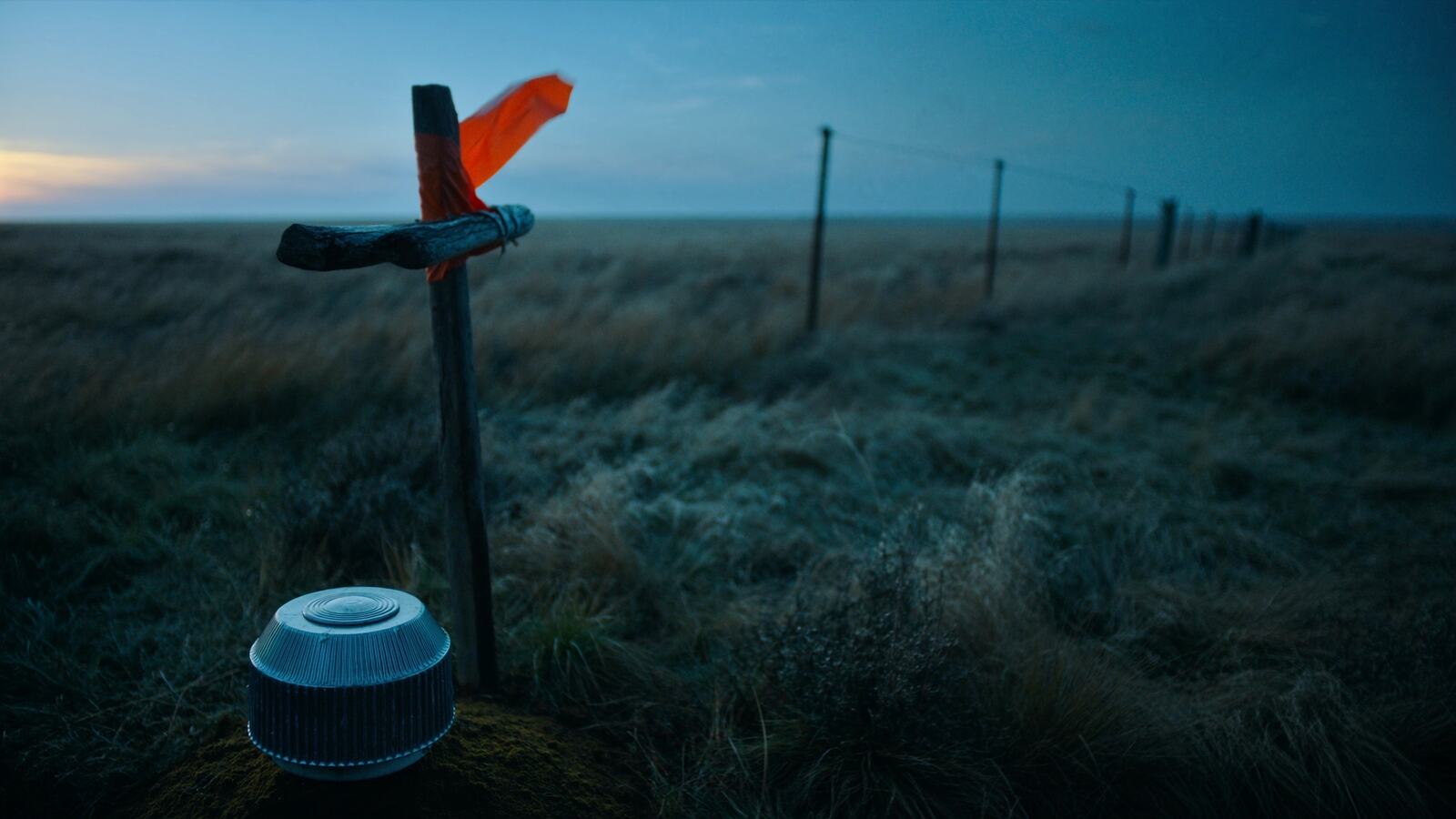 An aluminium-capped survey monument set flush in prairie soil at dawn beside a wooden stake wrapped in bright orange flagging tape, with a straight fence line running to the horizon.